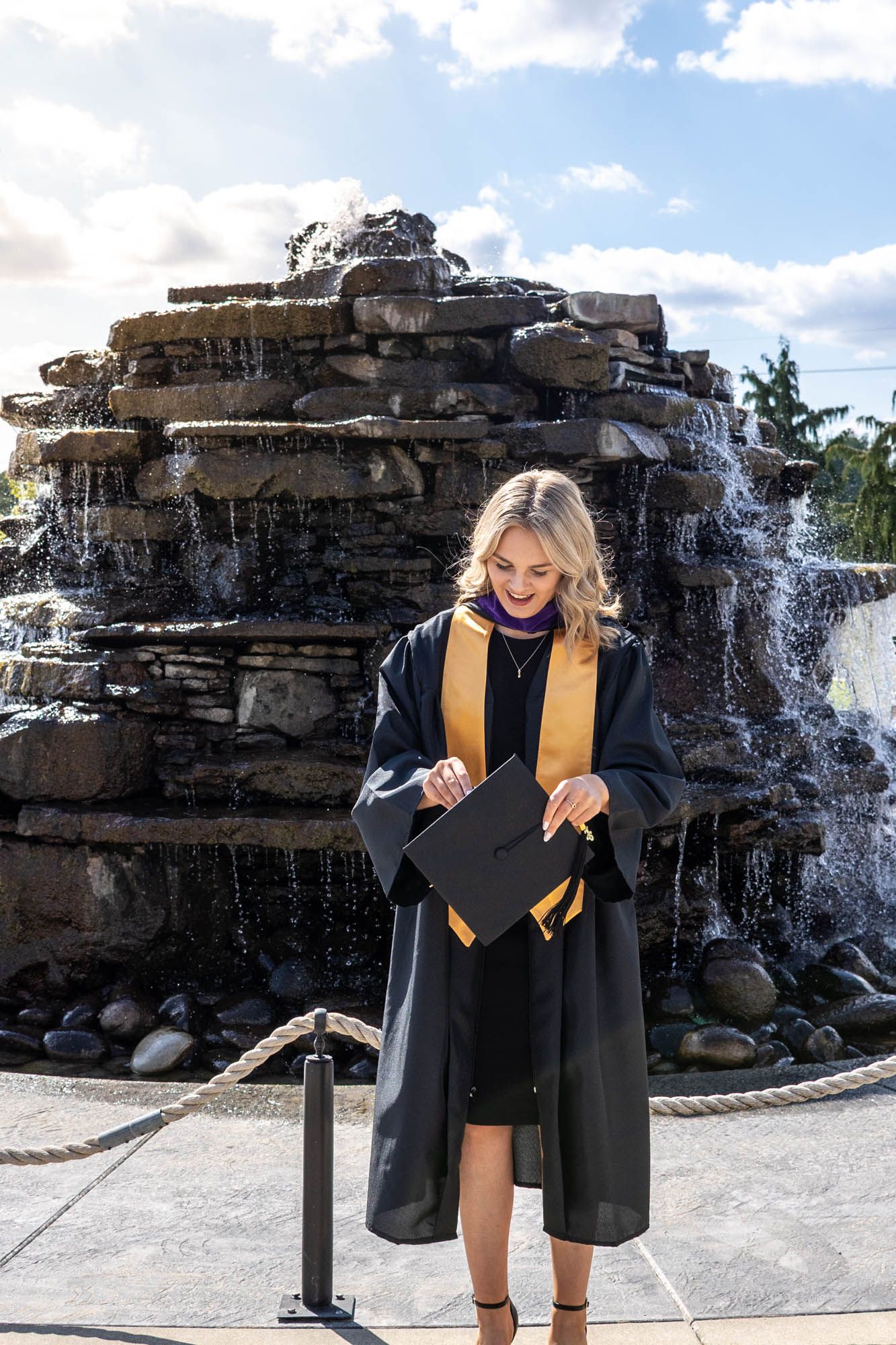 Smiling blonde woman in black graduation gown with gold stole holding cap in front of stone waterfall.
