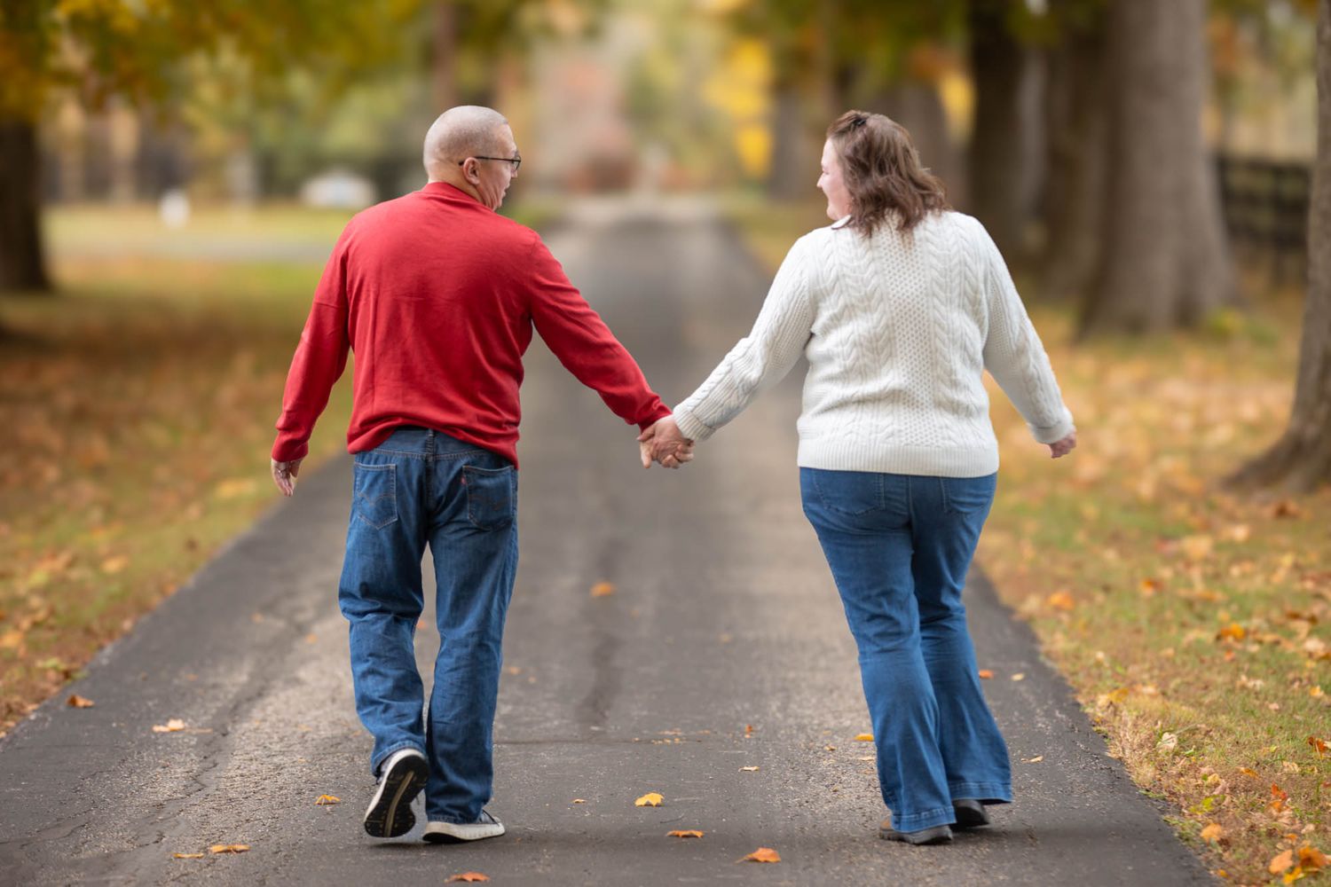 Couple walks hand in hand down a tree-lined road surrounded by fall foliage from behind.