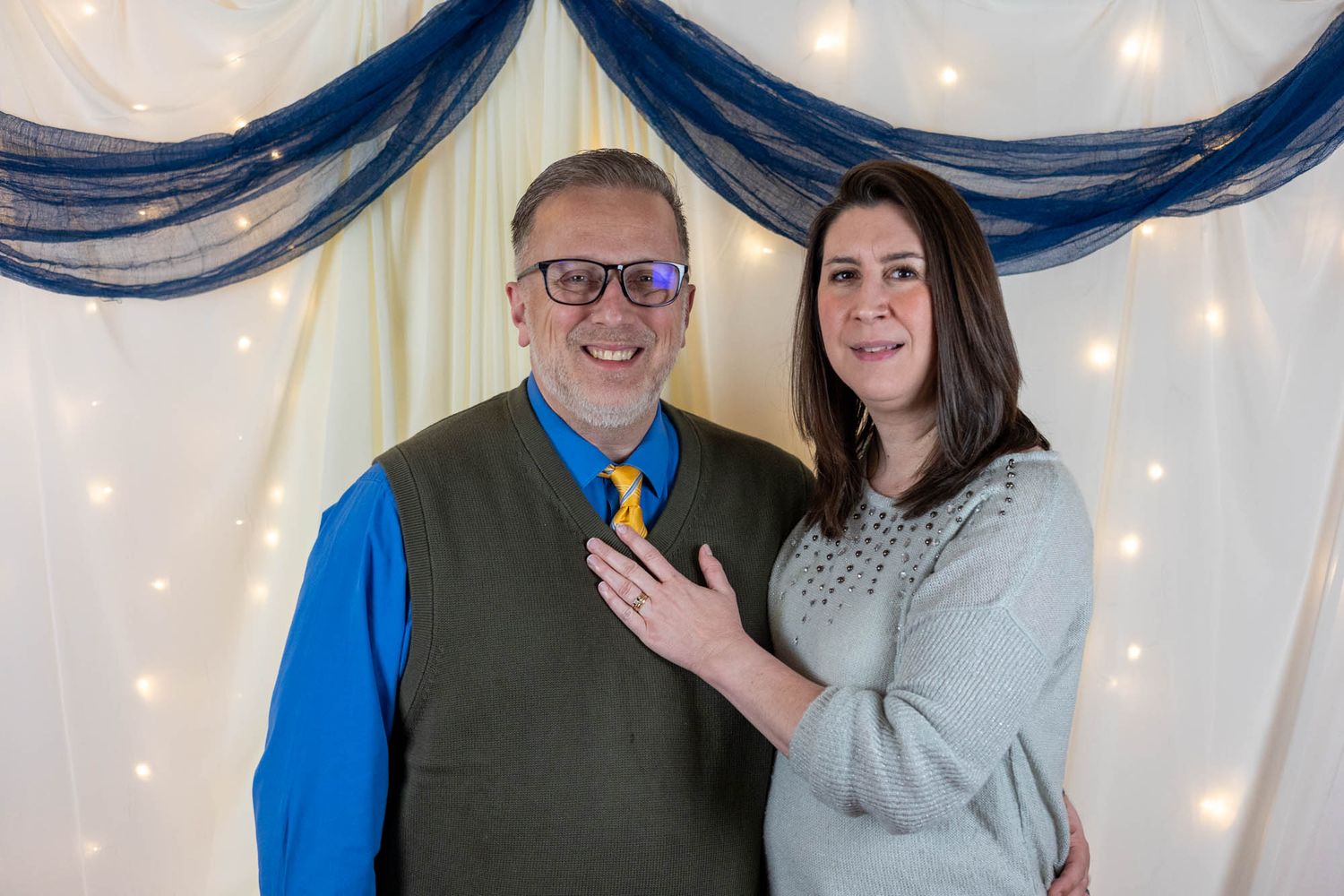 A cheerful couple poses together, the woman resting her hand on the mans chest before a decorated backdrop.