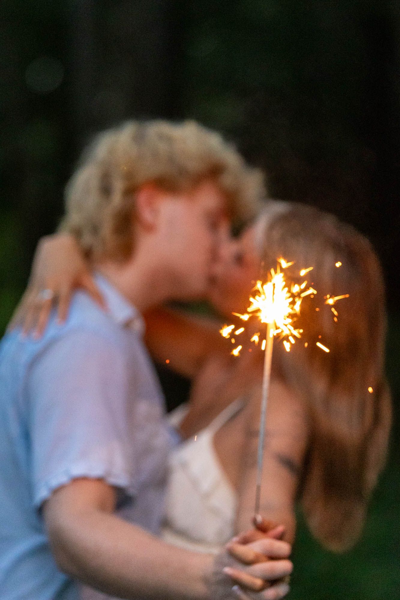A couple kissing outdoors at dusk while holding a glowing sparkler, captured in a romantic blurred bokeh style.