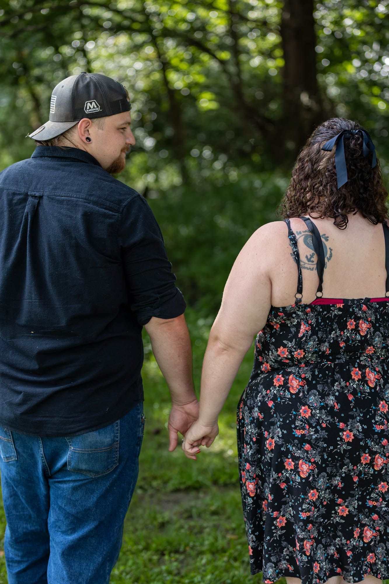 A couple holds hands while walking through a lush green forest, man in black shirt and cap, woman in floral dress.