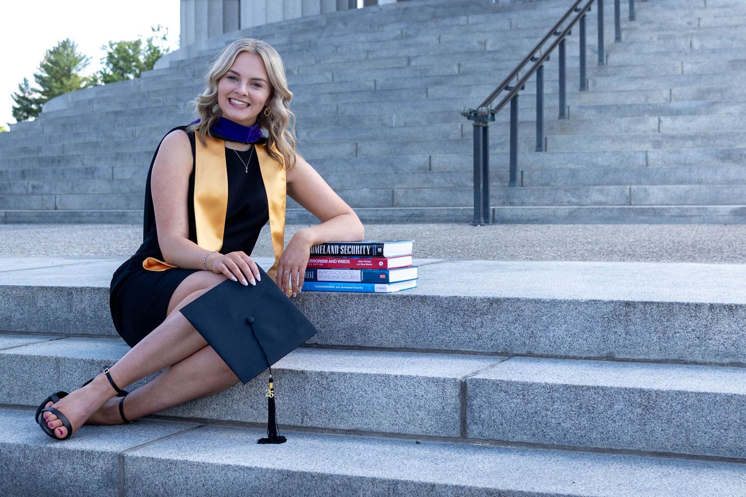 Smiling graduate in cap and gown colors sits on stone steps with stacked books, celebrating academic achievement outdoors.
