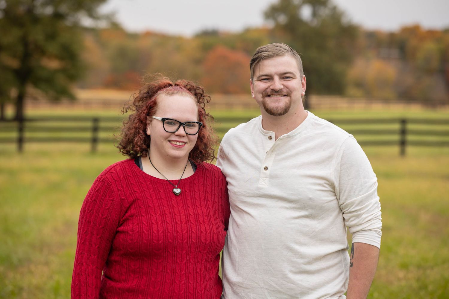 Smiling couple posing outdoors in autumn field, woman in red sweater and man in white henley shirt.