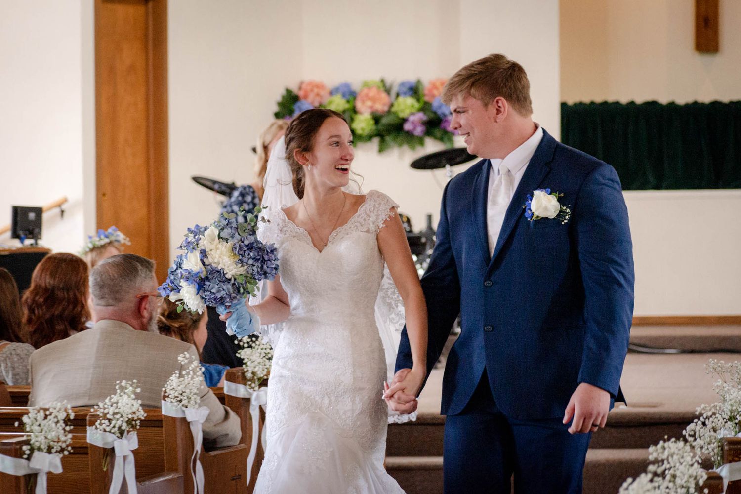 Happy newlywed couple holding hands and smiling while walking down church aisle after wedding ceremony.