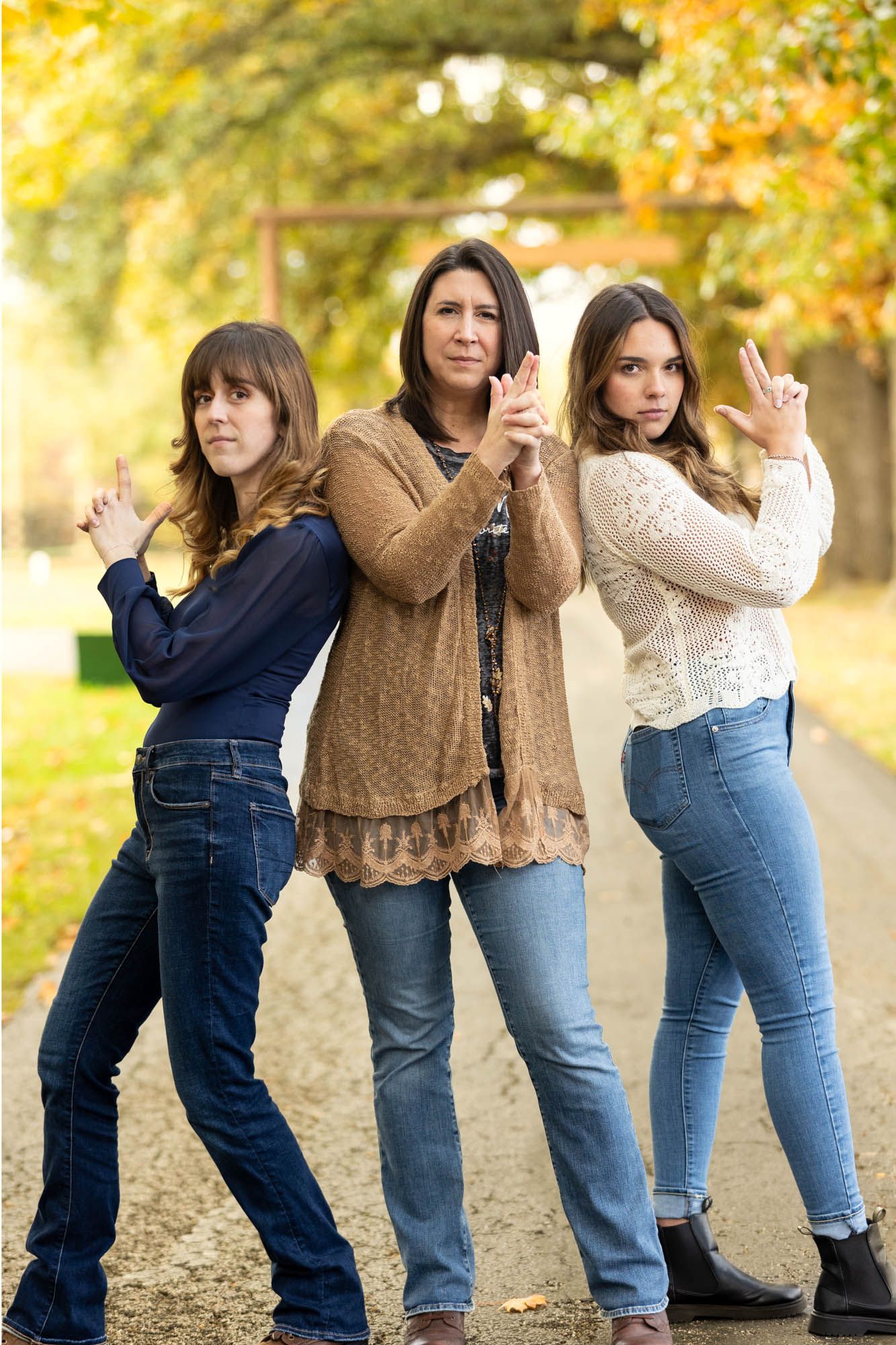 Three women strike fierce action poses on a scenic autumn tree-lined path in casual outfits.