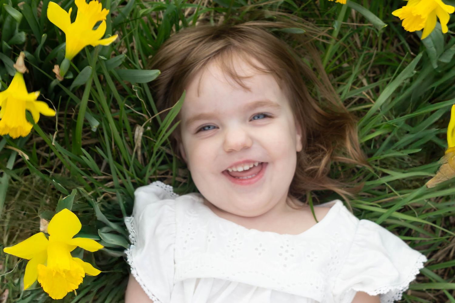 Happy young girl in a white dress lying in green grass surrounded by bright yellow daffodil flowers.