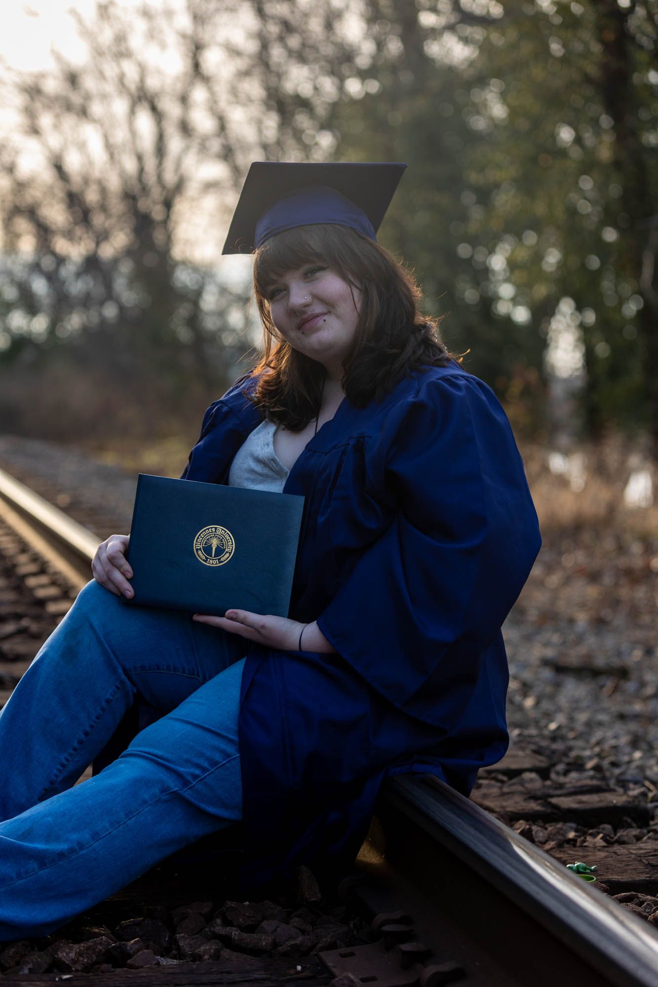 Graduate in blue cap and gown sits on railroad tracks holding a diploma in a scenic outdoor photo.