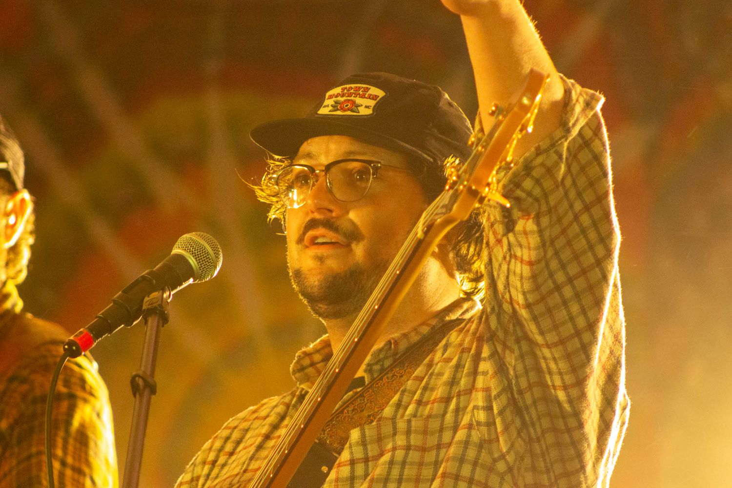 Bearded guitarist in cap raises hand while performing on stage under warm yellow lights.