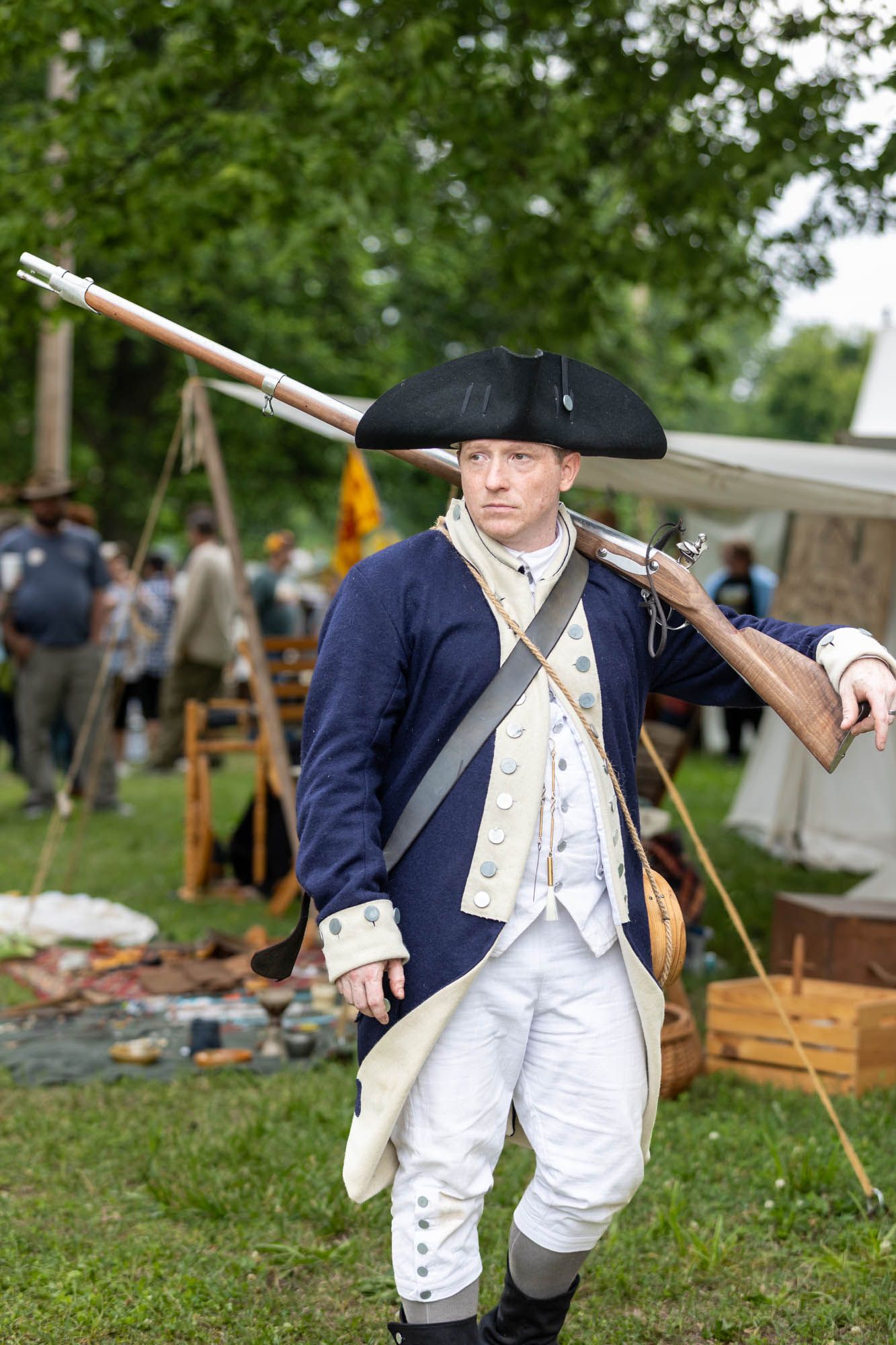A male reenactor in colonial Continental Army uniform carries a musket rifle at an outdoor historical event.