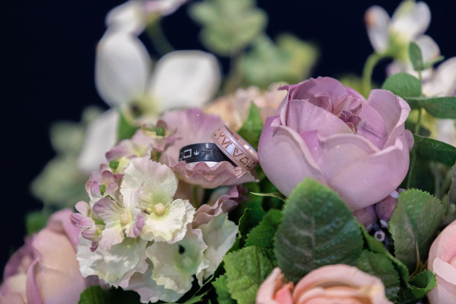 Silver wedding rings resting on a bouquet of pink roses and white hydrangeas against a dark background.