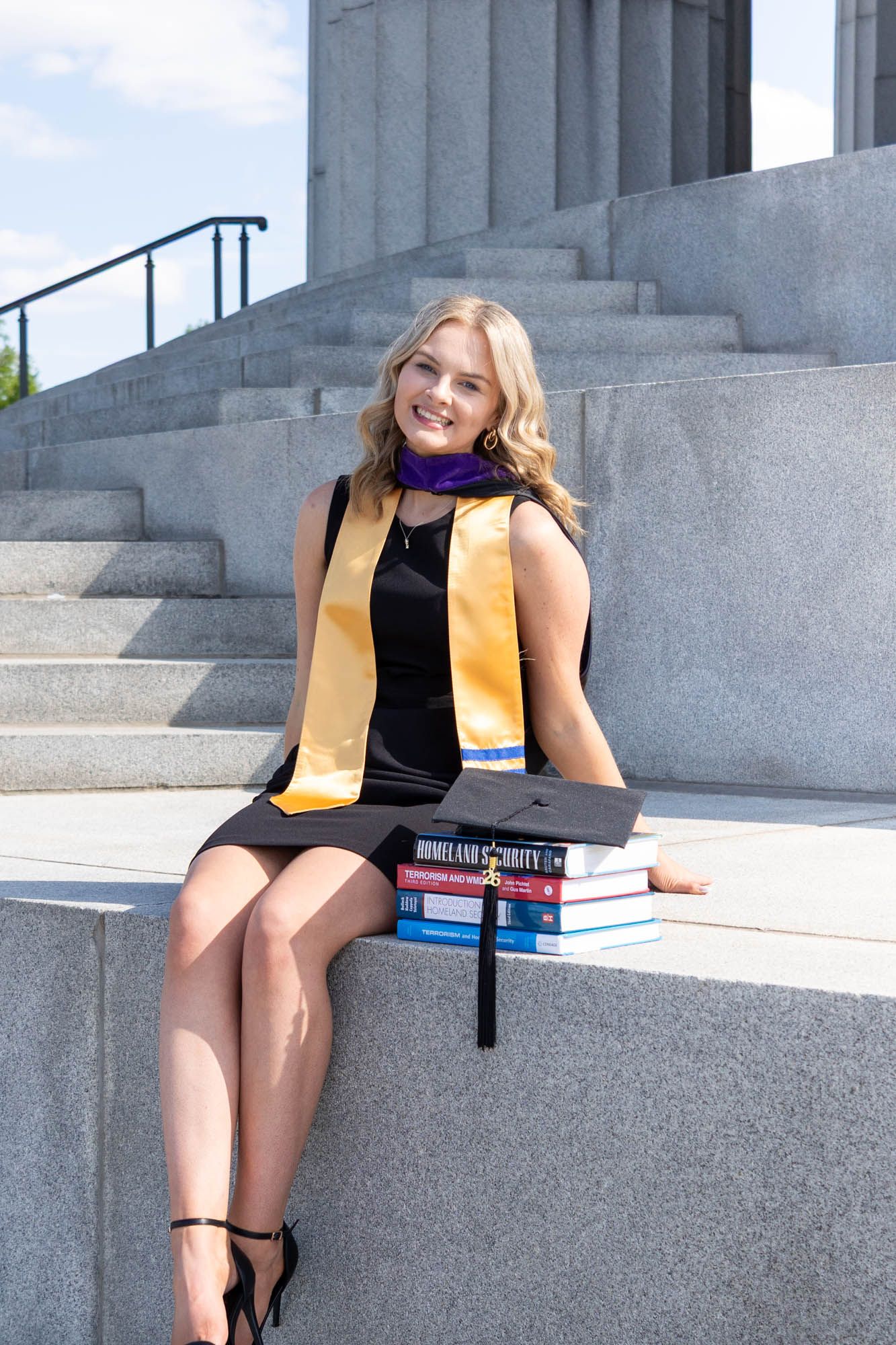 Smiling graduate in black dress with gold and purple stole sits on stone steps with graduation cap and textbooks.