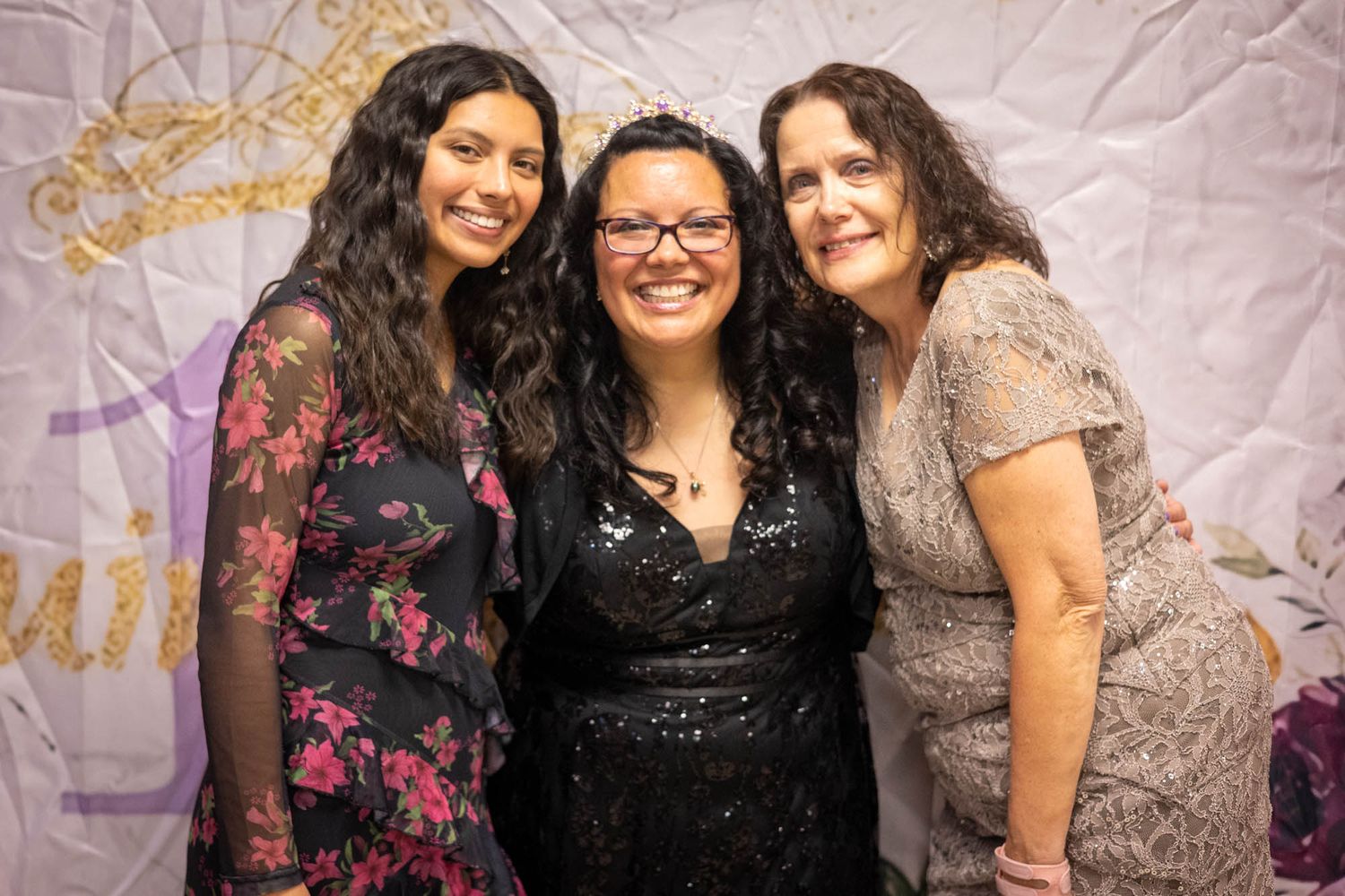Three women smile together in front of a decorative backdrop at a quinceañera reception celebration.