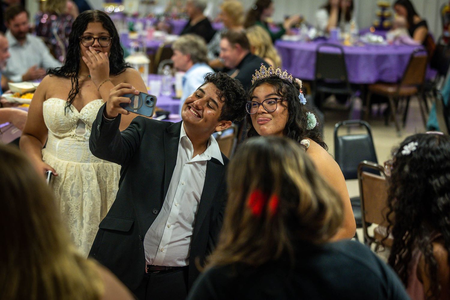 Guests take a selfie at a quinceañera reception, with a girl wearing a tiara posing with friends.