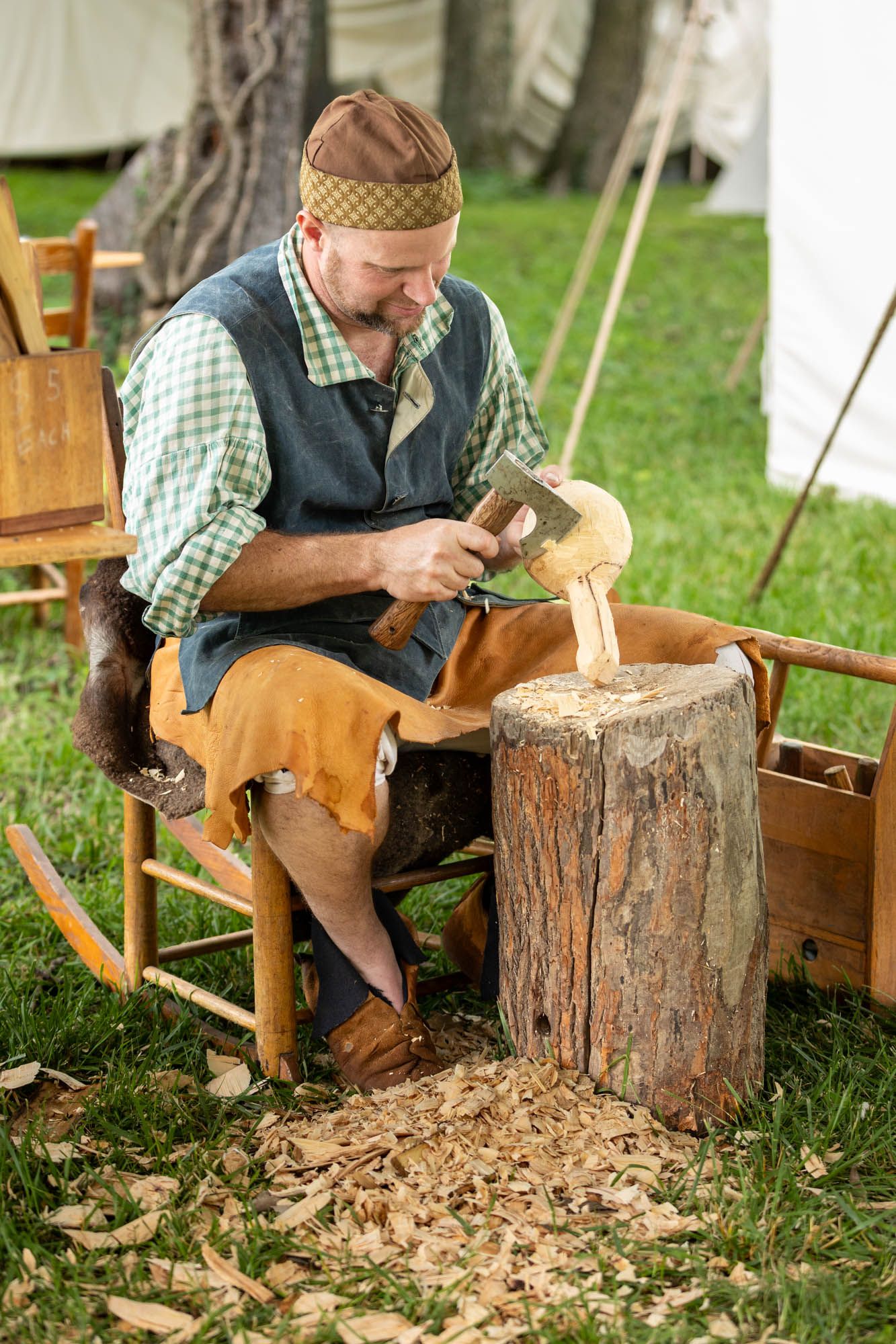Craftsman in historical costume carving wood with an axe on a tree stump at an outdoor heritage festival.