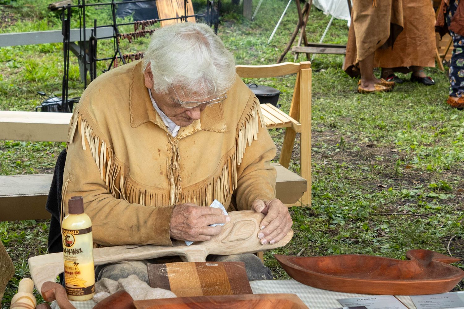 Elderly craftsman in fringed leather jacket carefully carving wood at an outdoor historical demonstration event.