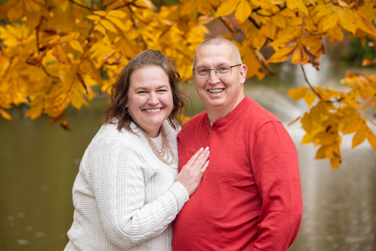 Smiling couple poses together under golden yellow autumn leaves near a peaceful pond.