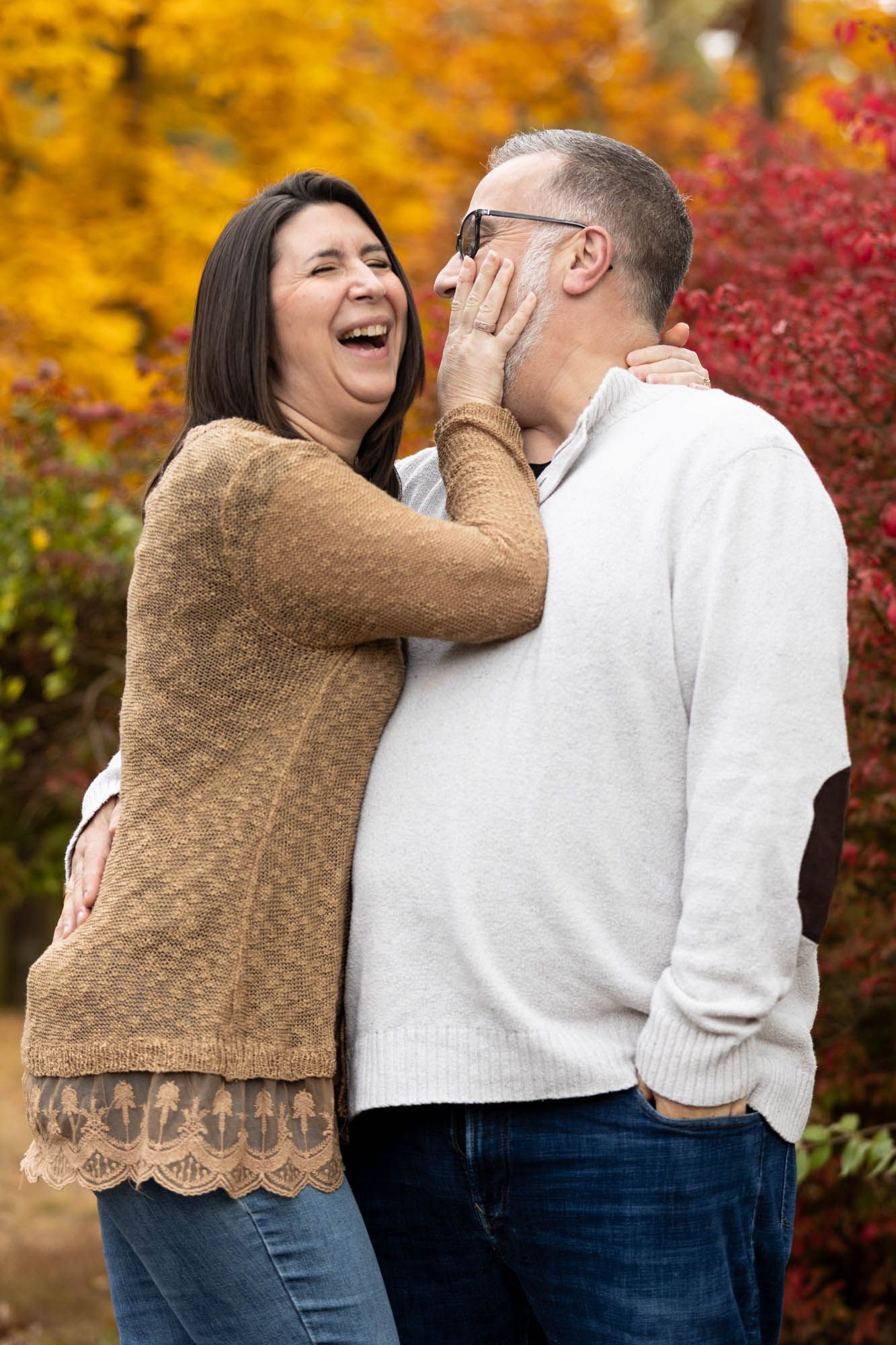 Laughing couple in casual sweaters share a joyful moment outdoors among colorful autumn foliage.