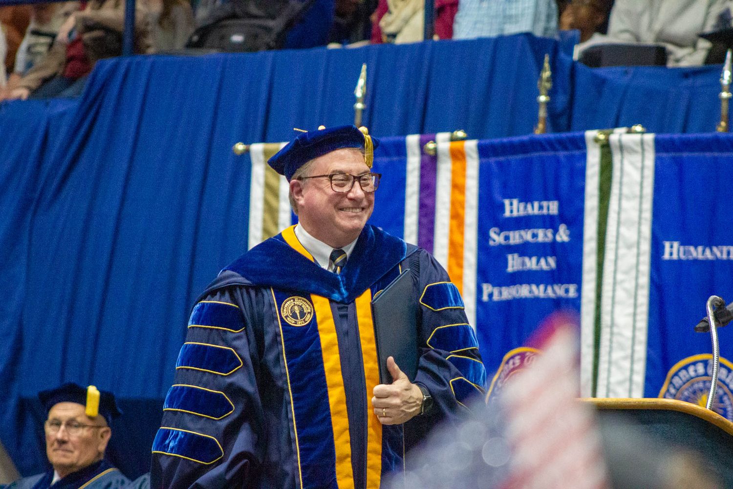 Smiling university official in blue and gold academic regalia walks during graduation ceremony.