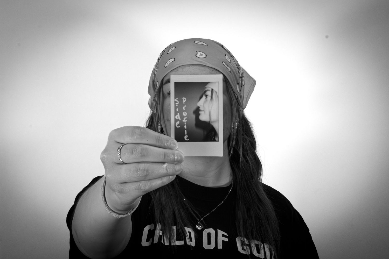 Woman in a Child of God shirt holding a Polaroid photo over her face in a black and white portrait.