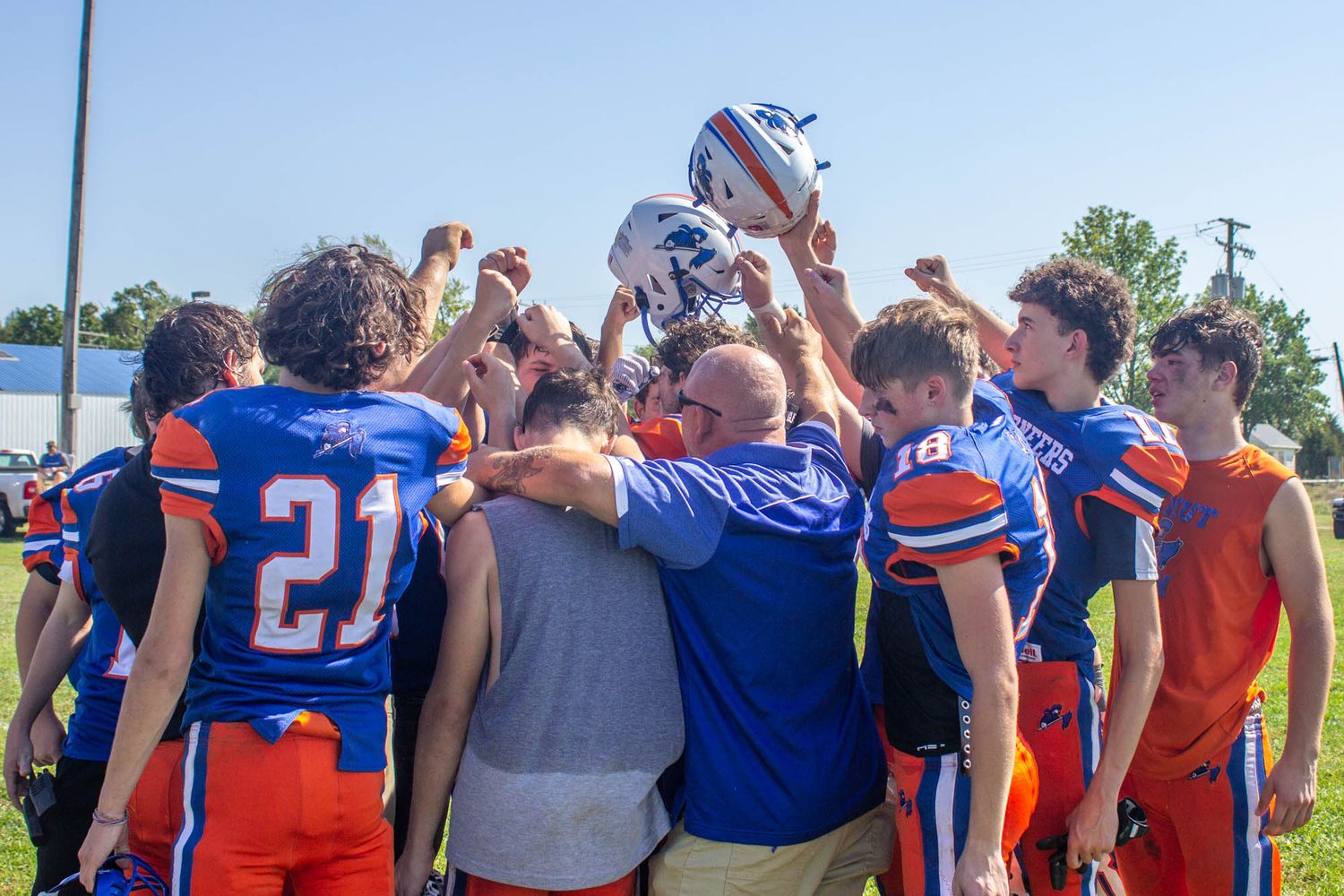 Football team huddles together celebrating victory, players in blue and orange uniforms raising helmet.