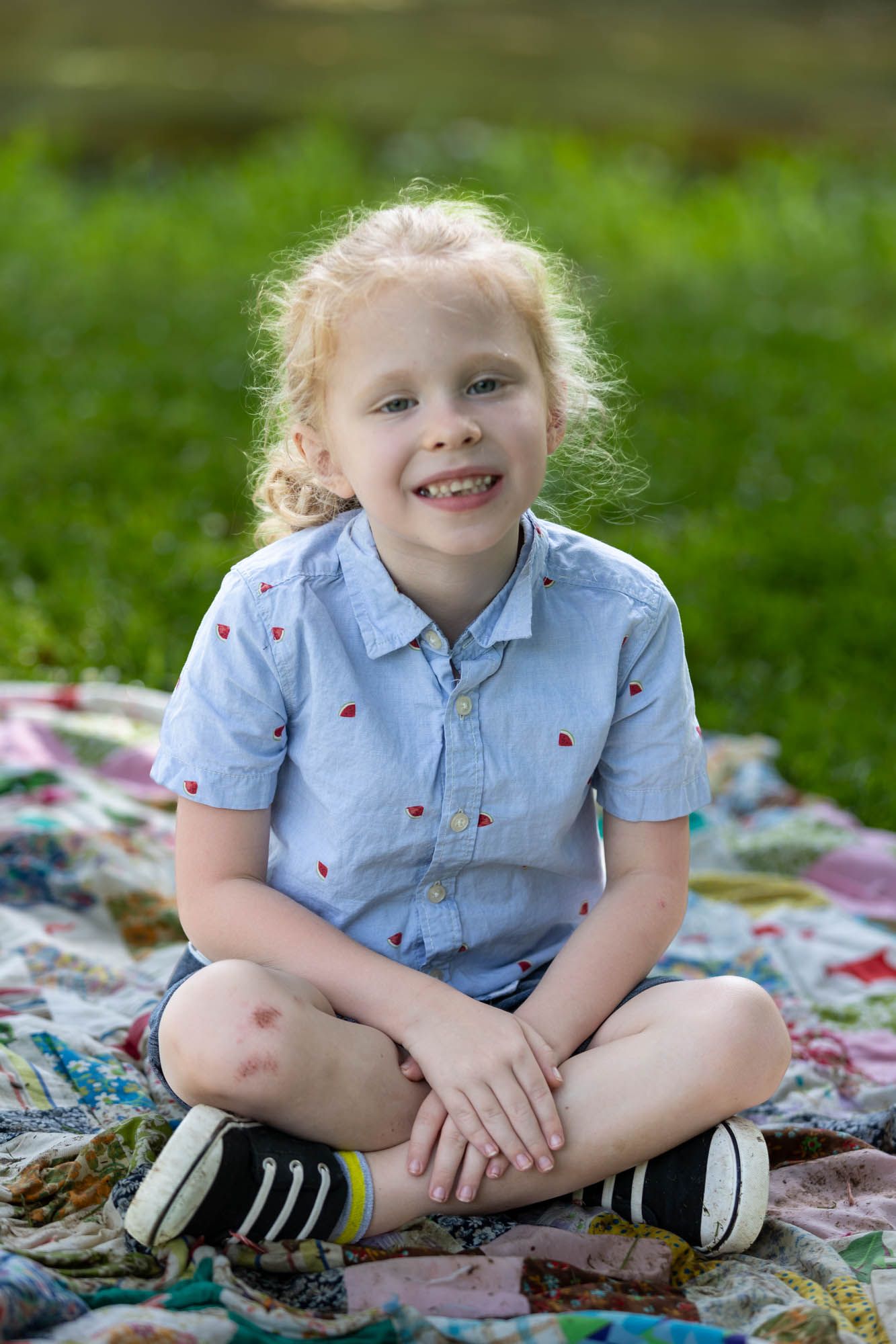 Smiling blonde girl sitting cross-legged on a colorful quilt blanket outdoors on green grass.