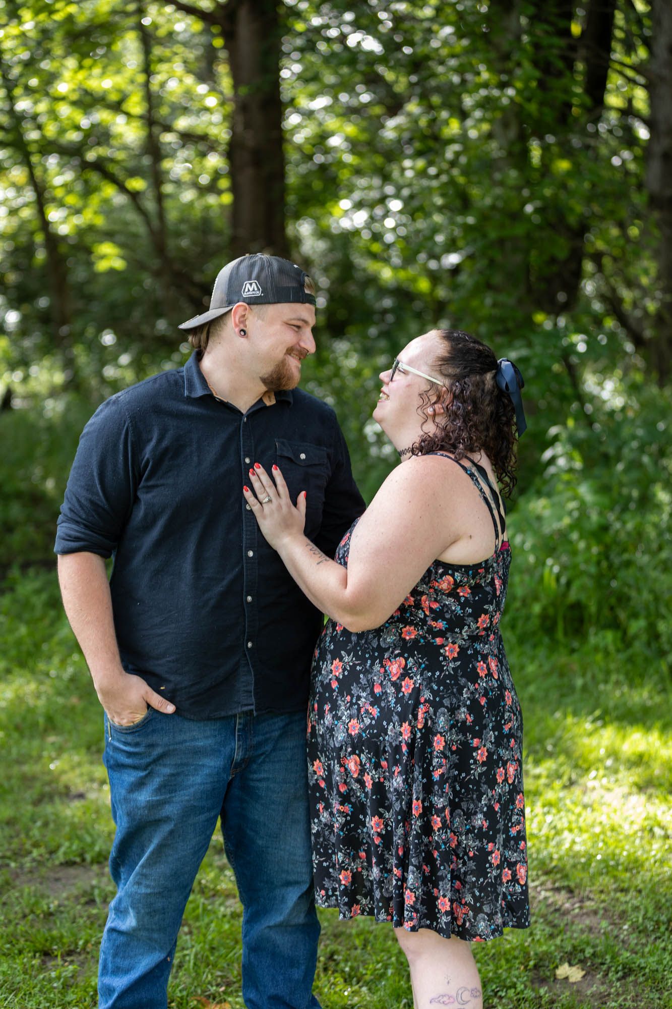 Couple in a lush green park, woman in floral dress places hand on man's chest as they gaze at each other.