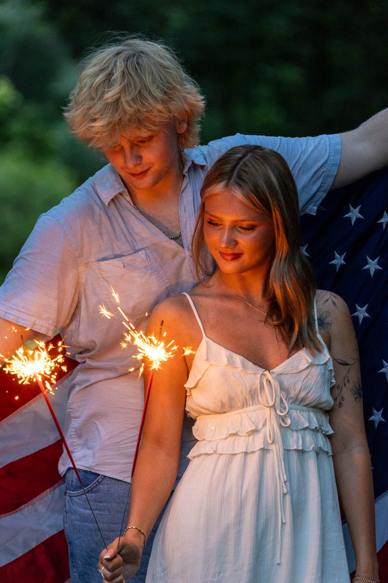 Young couple holding sparklers with an American flag on Fourth of July celebration outdoors at dusk.