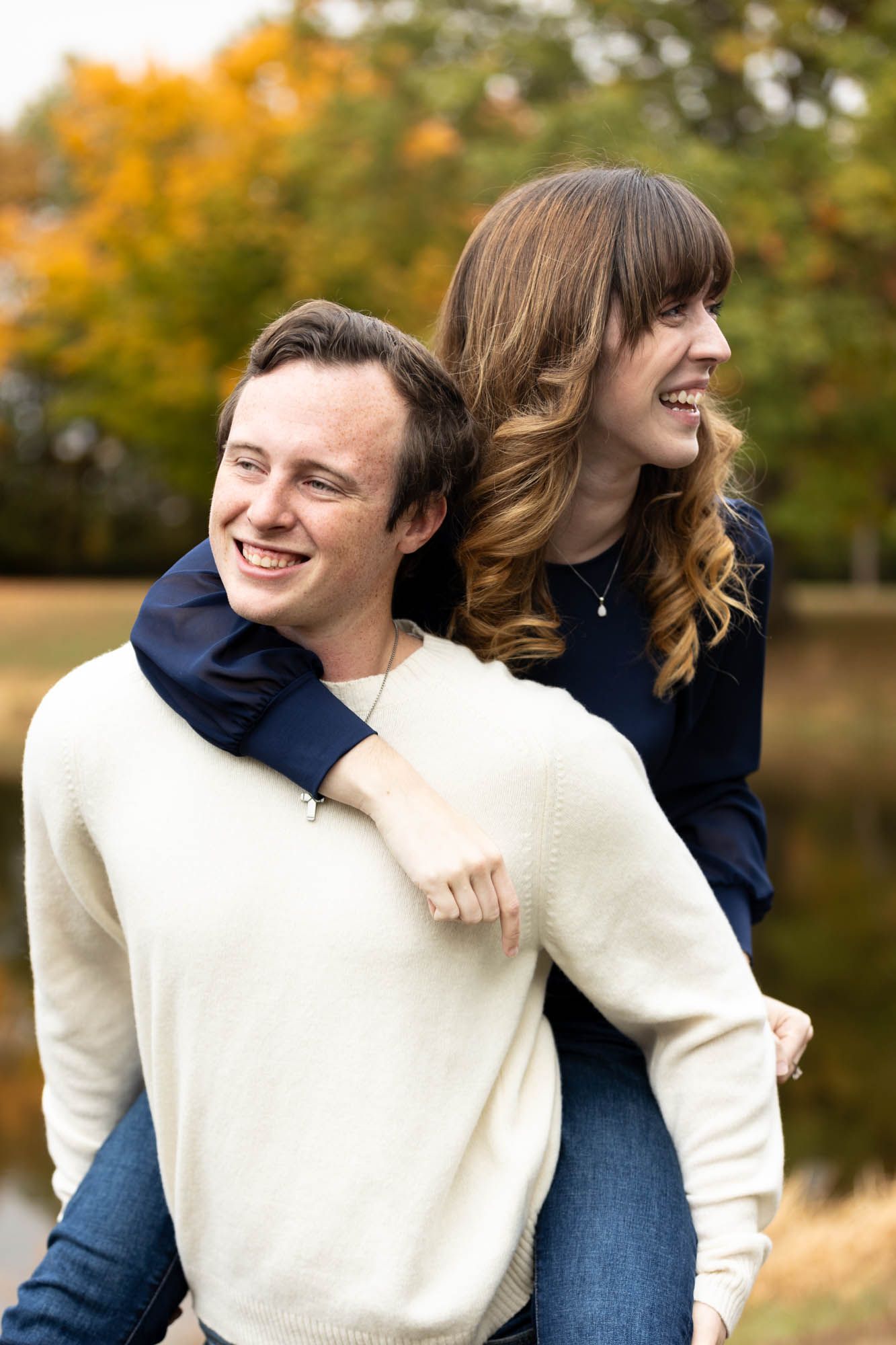 Man gives woman a piggyback ride while both laugh joyfully during autumn engagement photos.