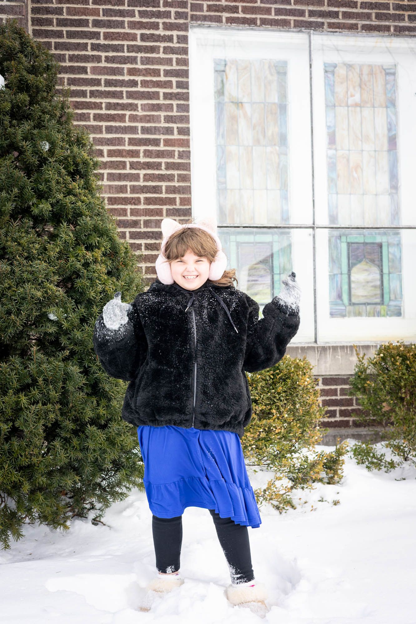 Young girl in black fur jacket and blue skirt wearing pink earmuffs playing in snow outside.