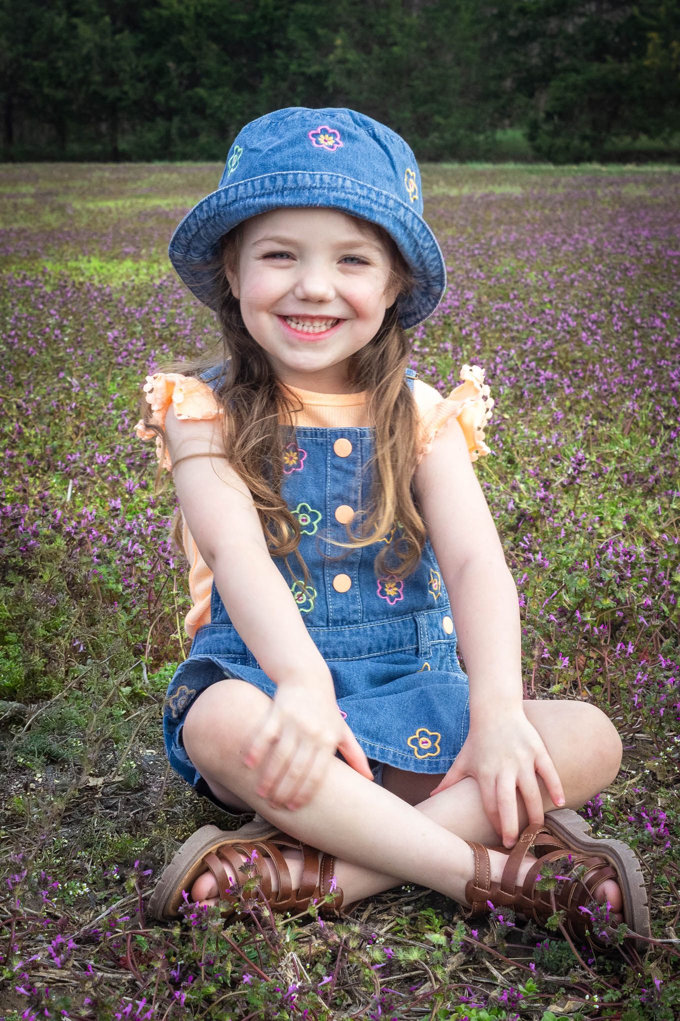 A smiling young girl in denim overalls and blue bucket hat sits cross-legged in a purple wildflower meadow.