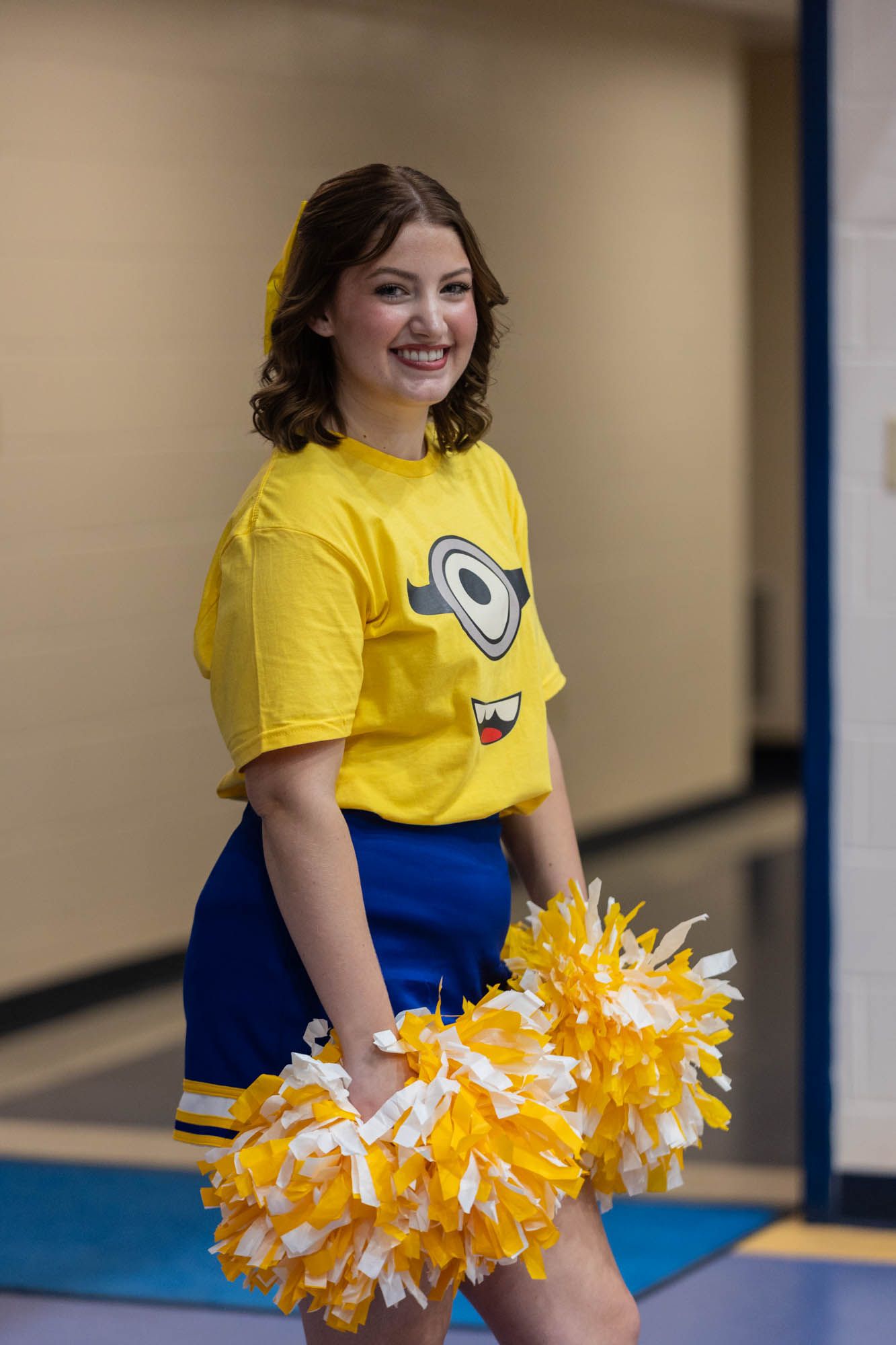 Cheerleader in yellow Minion t-shirt and blue skirt smiles holding gold and white pom-poms.