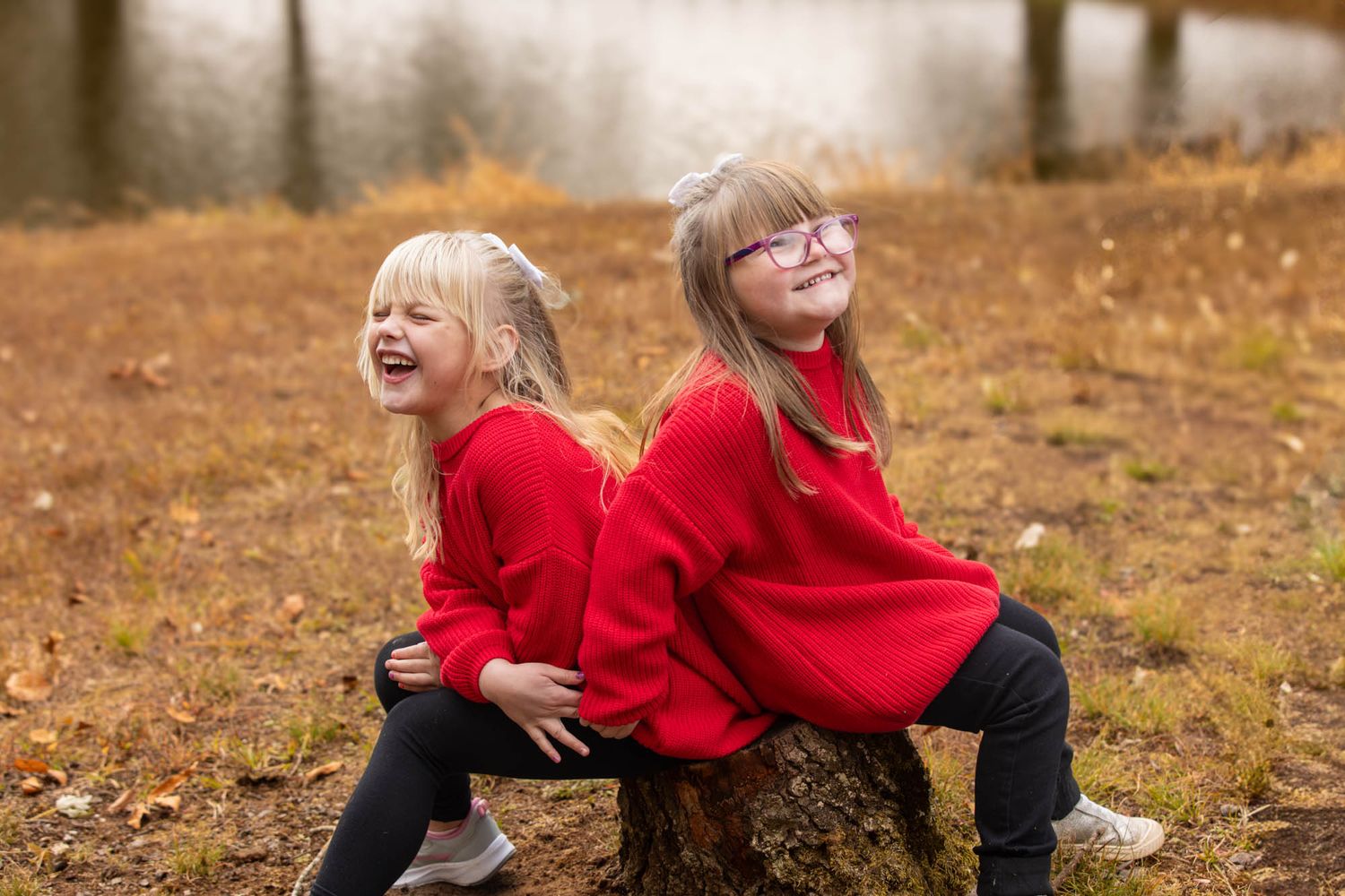 Two young girls in matching red sweaters laugh while sitting back-to-back on a tree stump in an autumn park.