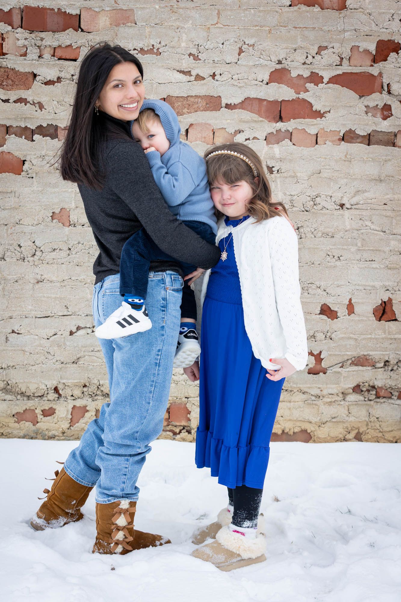 Mother holding young son while daughter in blue dress stands beside her against a snowy brick wall.