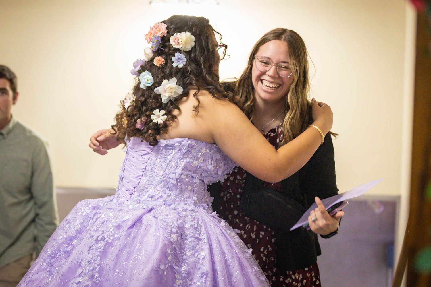 A woman smiling and hugging a girl in a purple quinceañera gown with floral hair accessories at a celebration.