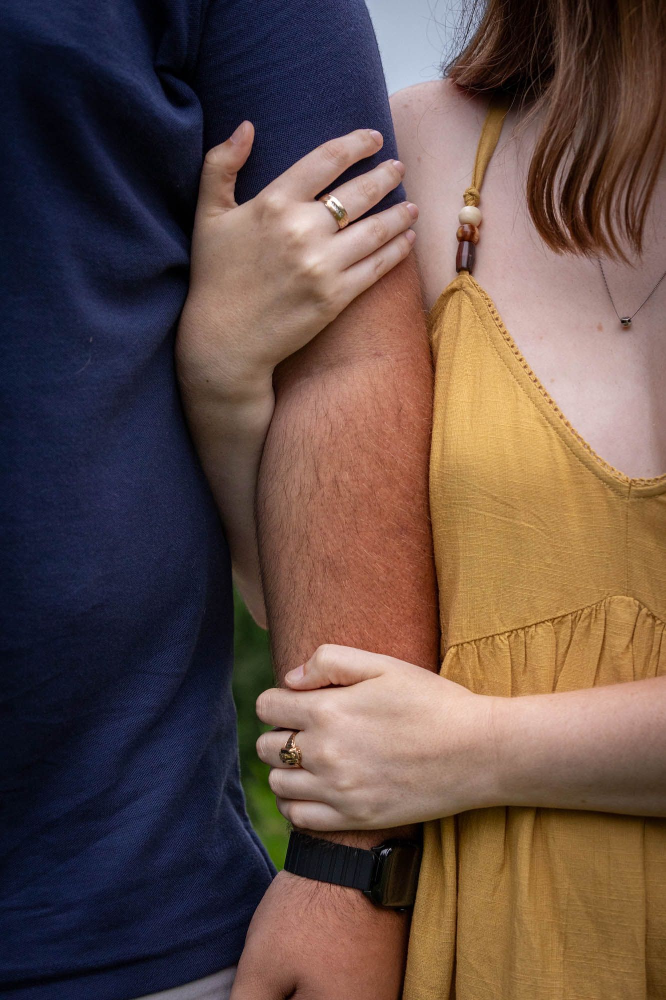 Close-up of a couple holding hands, wearing matching gold rings, she in a yellow dress, he in a navy shirt.