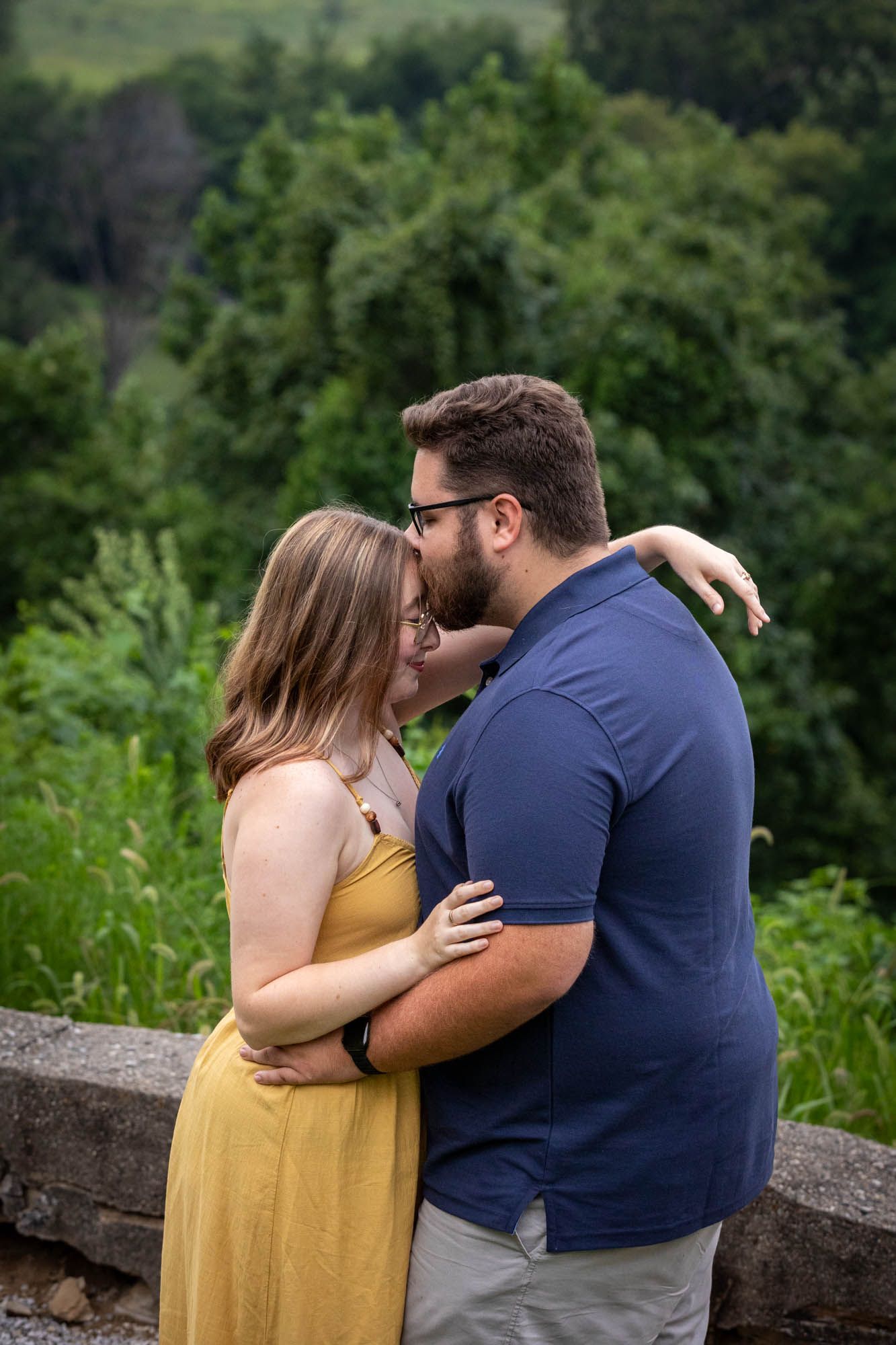 A couple embraces and kisses outdoors, surrounded by lush green trees, woman in yellow dress, man in navy shirt.