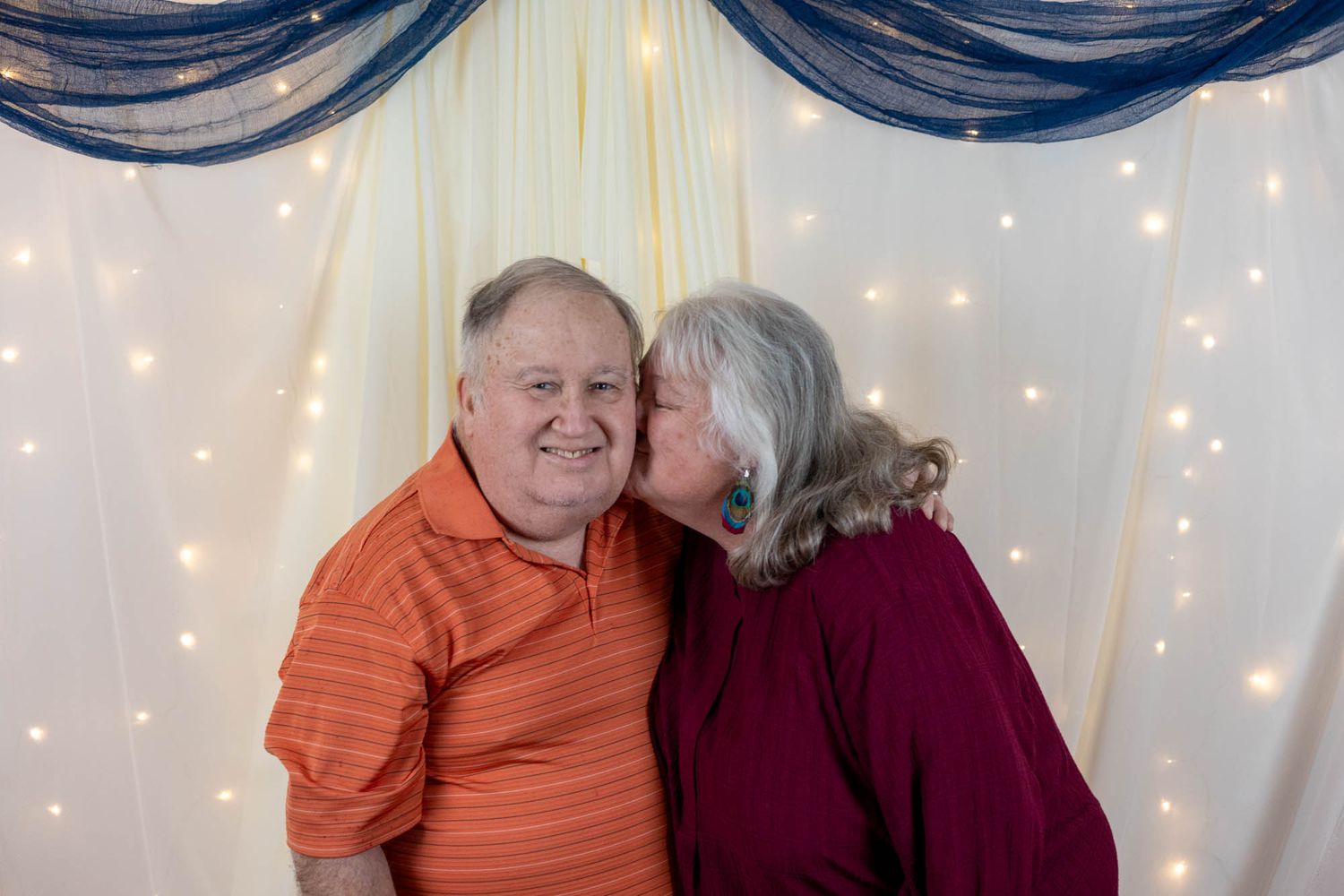 An elderly couple shares a sweet moment as the woman kisses the man on the cheek before a decorated backdrop.