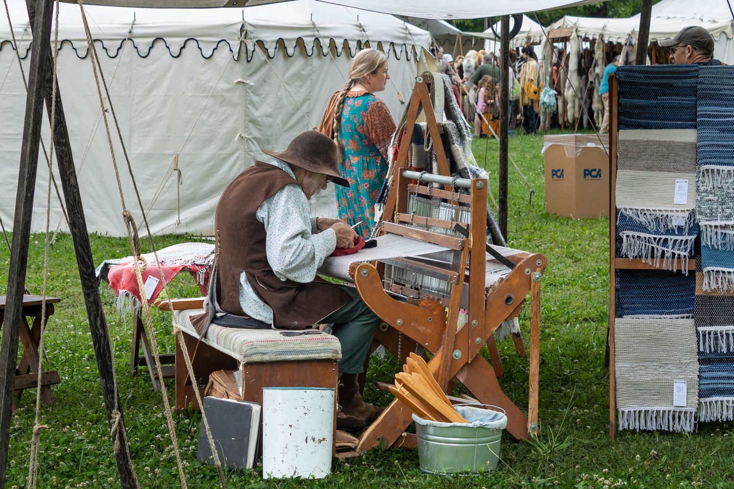 Person in period costume weaving on a wooden loom at an outdoor historical fair with tents in background.