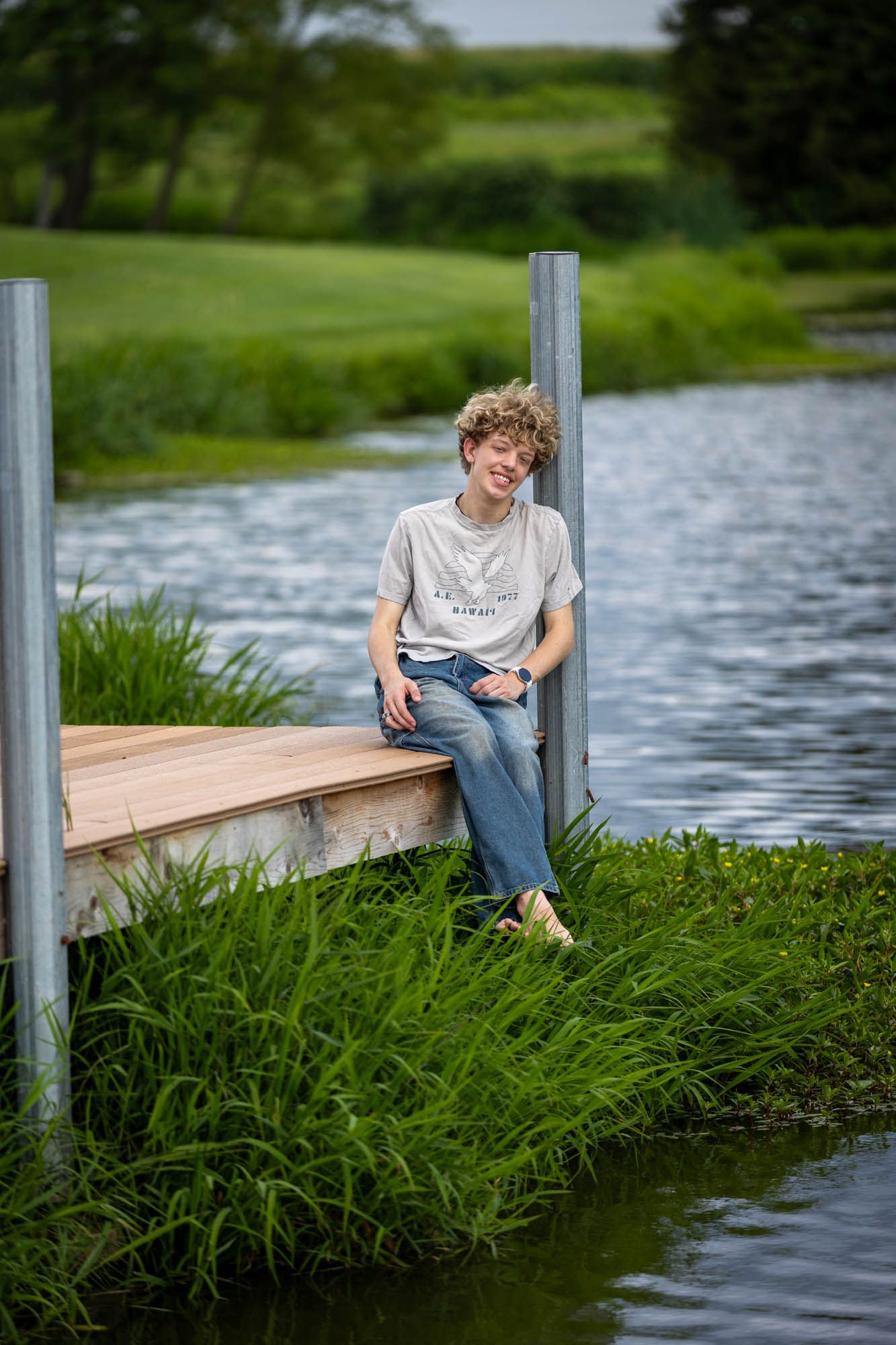 Teen with curly hair sits on a wooden dock by a calm pond, wearing jeans and a gray tee, surrounded by lush greenery.