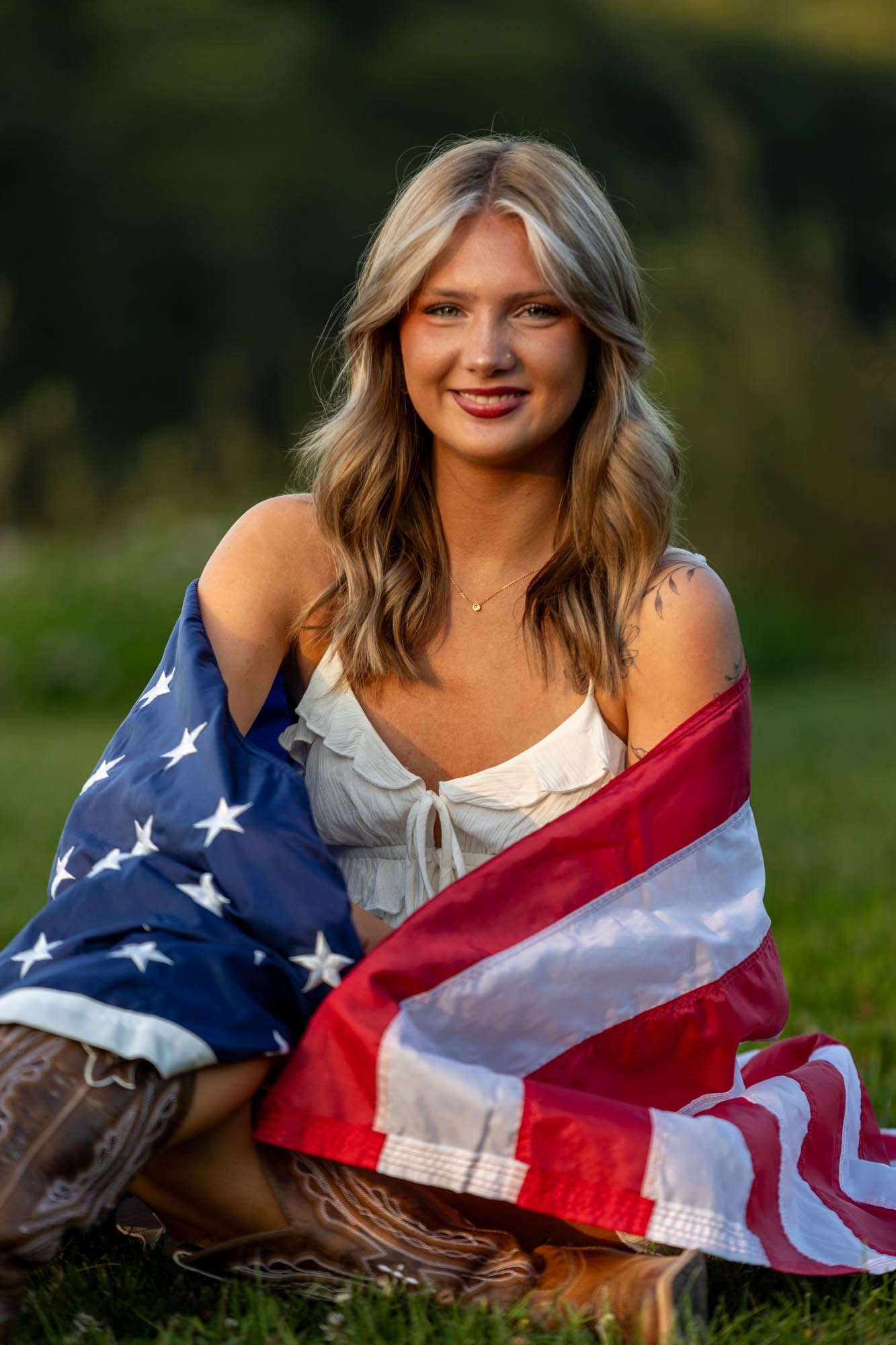 Young blonde woman wrapped in American flag sitting on grass, smiling outdoors in golden hour light.