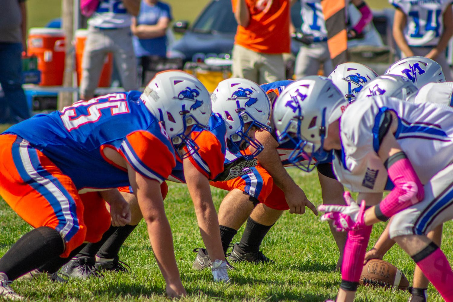 High school football players in blue and orange uniforms line up against opponents at the line of scrimmage.