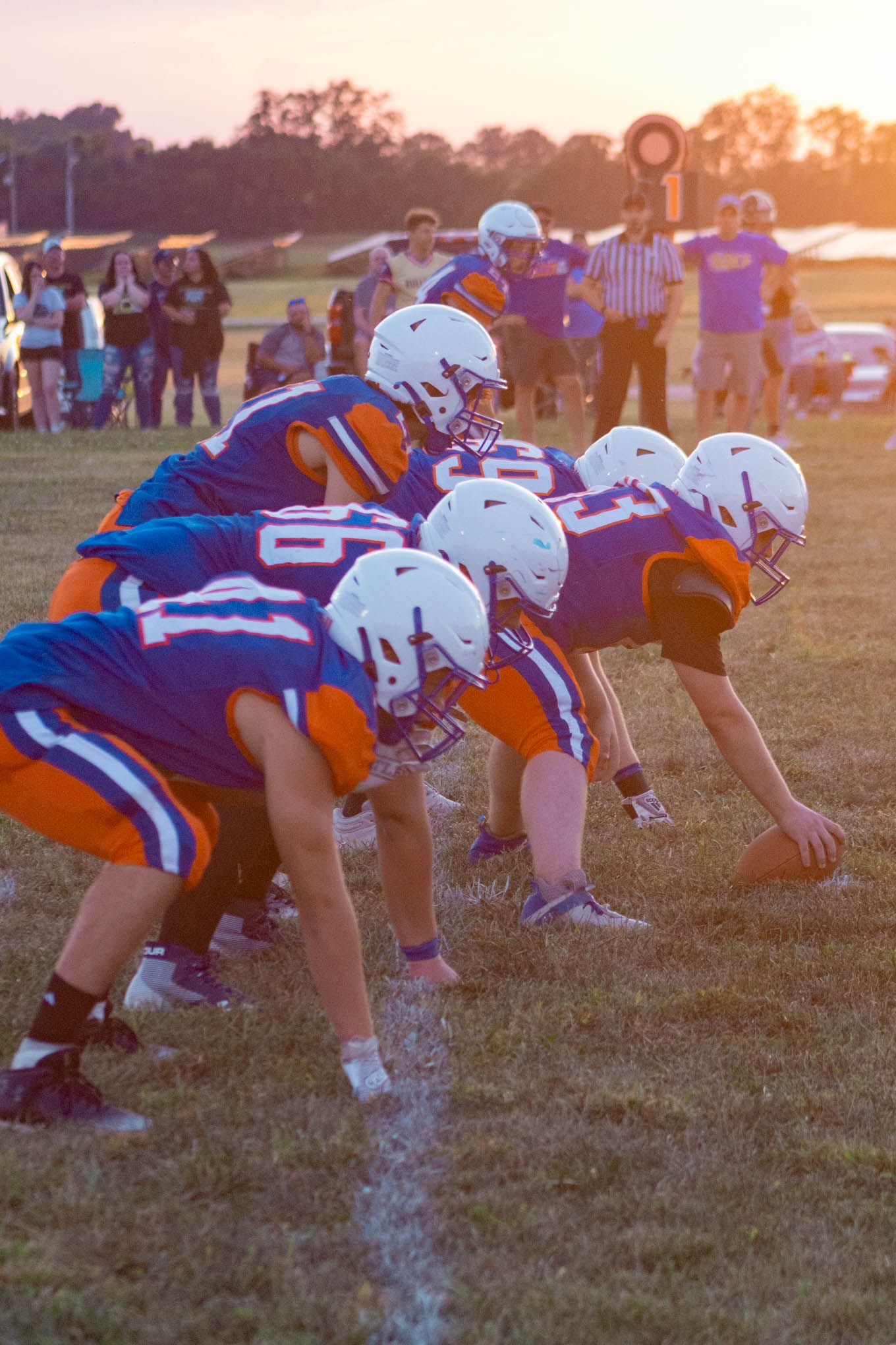 Youth football team in blue and orange uniforms lines up at scrimmage during a sunset game.