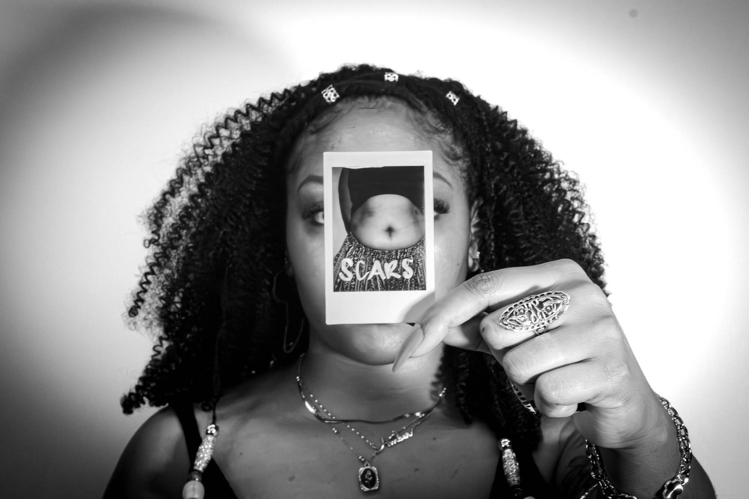 Black and white portrait of a woman with curly hair holding a Polaroid labeled SCARS over her face.