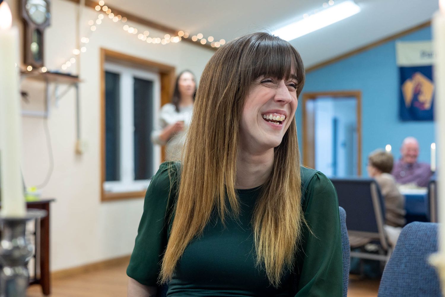 A young woman with long brown hair laughs joyfully while wearing a green dress at an indoor gathering.