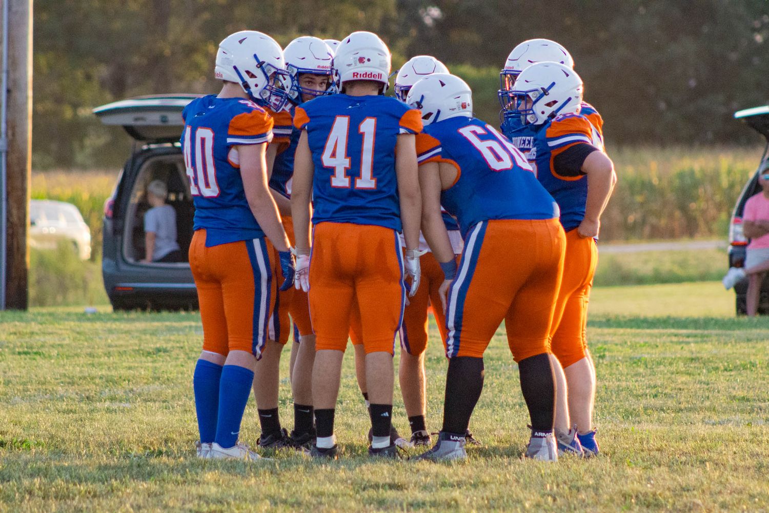 Youth football players in blue and orange uniforms huddle together on a green field during a game.