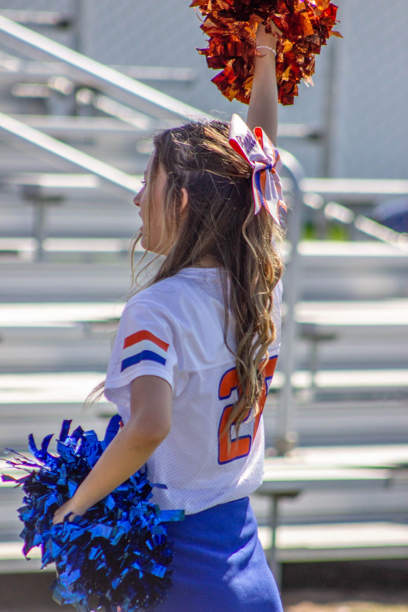 Cheerleader in blue and white uniform raises orange pom-pom near bleachers during a game.
