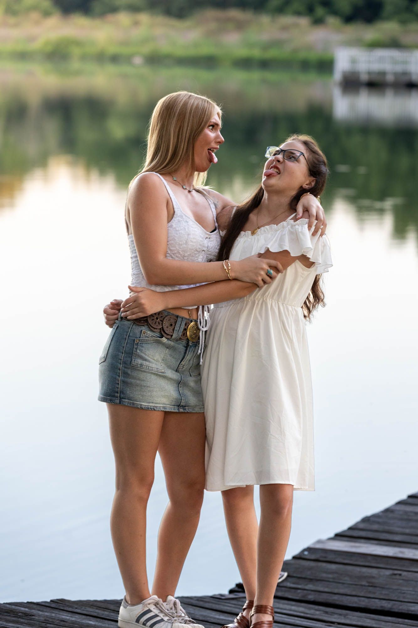 Two young women hugging and laughing together on a wooden dock by a calm lake on a summer day.