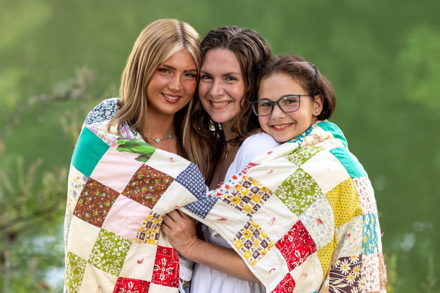 Three women wrapped in a colorful patchwork quilt smiling outdoors near lush green trees.
