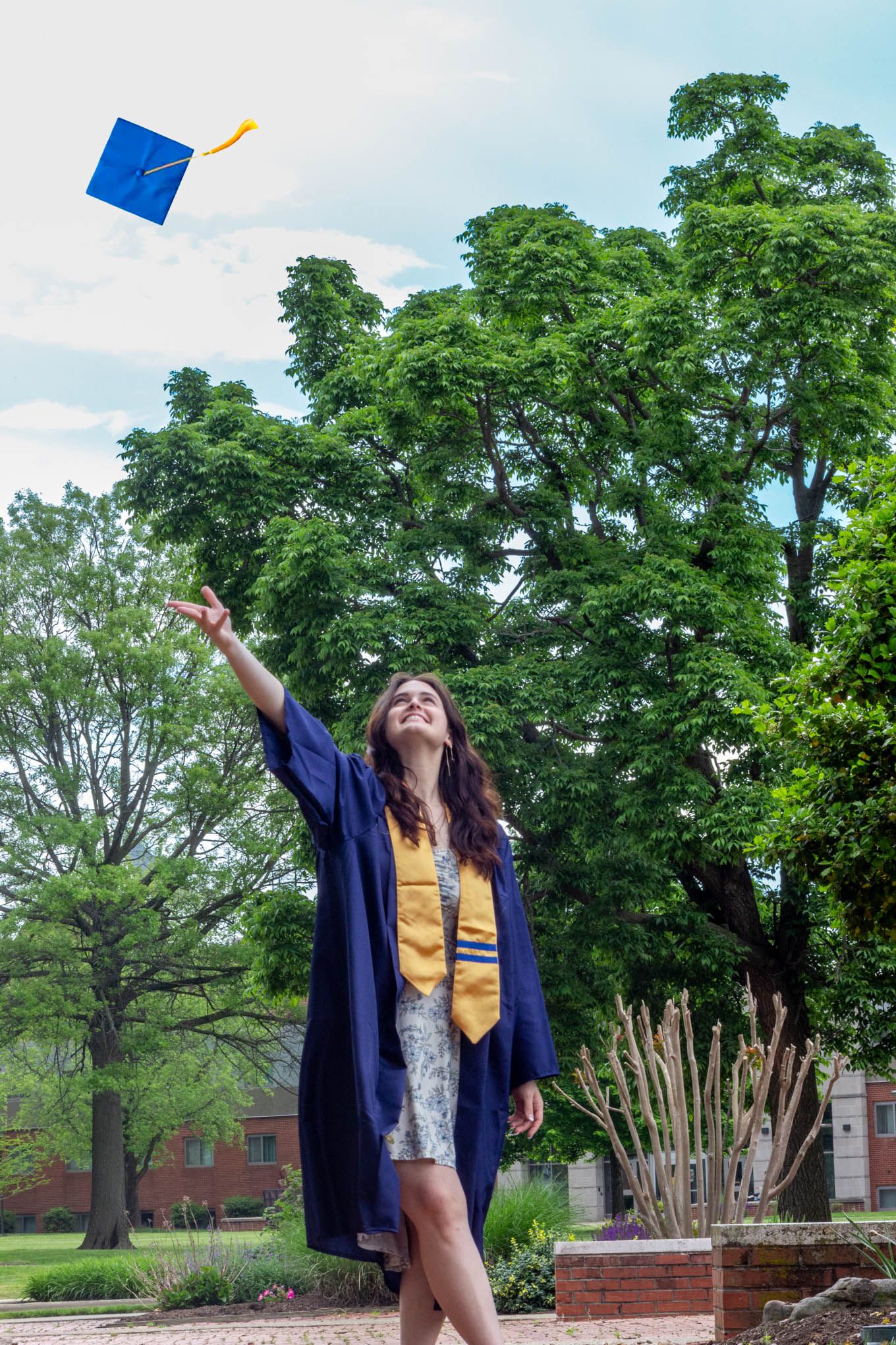 Joyful graduate in blue gown tosses her cap into the air in a lush green outdoor setting.