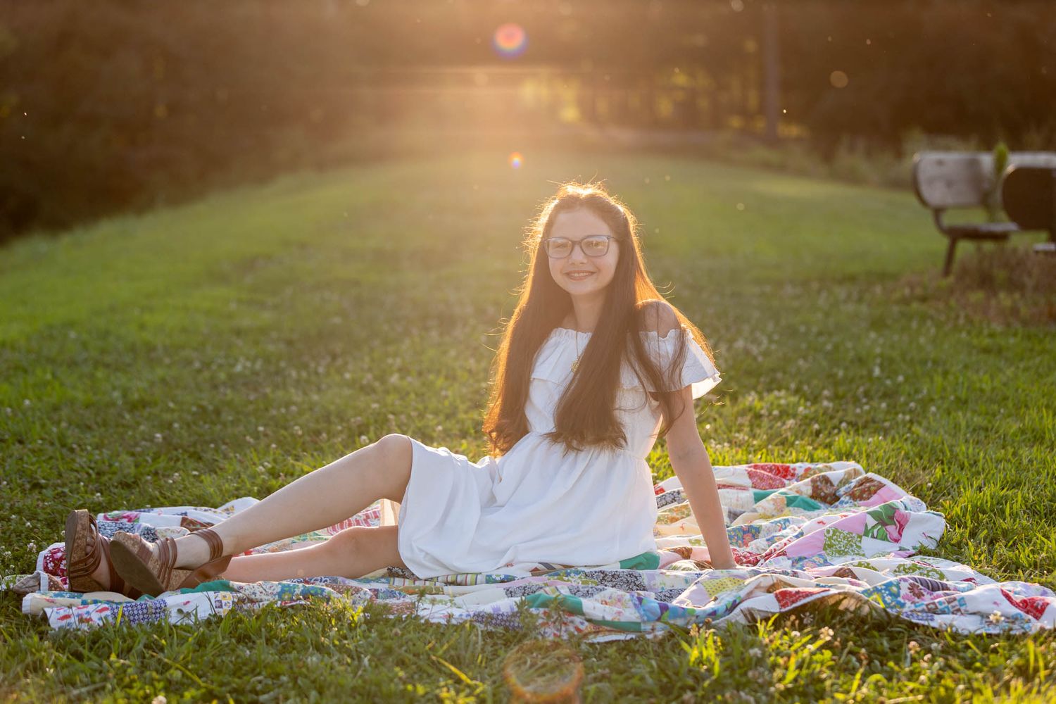 Young girl in white dress sitting on colorful quilt in sunny park, wearing glasses, smiling warmly at golden hour.